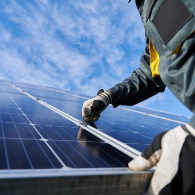 Close up of man technician in work gloves installing stand-alone photovoltaic solar panel system under beautiful blue sky with clouds. Concept of alternative energy and power sustainable resources.